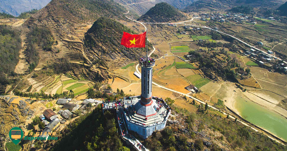 Lung Cu Flag Tower standing tall against a blue sky, with the Vietnamese flag fluttering in the wind atop the tower, surrounded by lush greenery